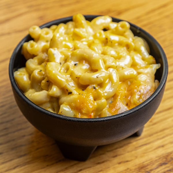 A black bowl filled with creamy, cheesy macaroni and cheese, topped with herbs, on a wooden table.