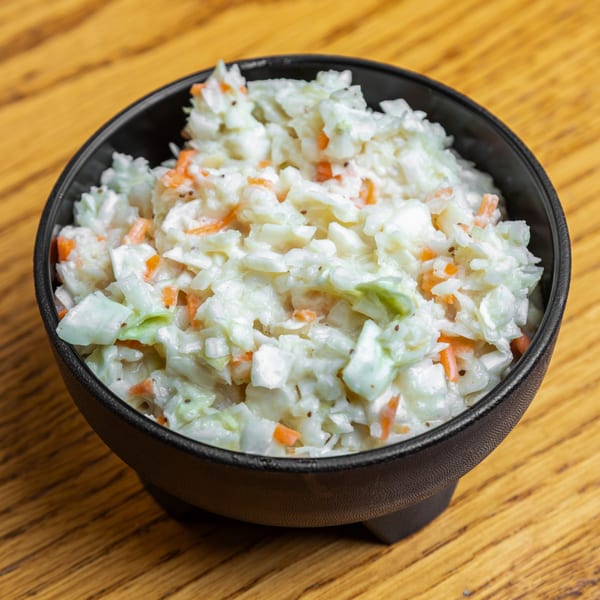 A black bowl filled with creamy coleslaw, featuring shredded cabbage and carrots, on a wooden table.