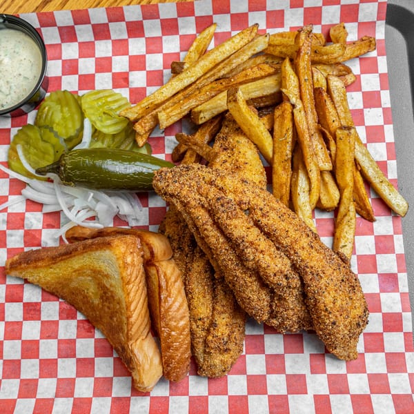 Fried catfish, french fries, toast, pickles, onions, jalapeño, and dipping sauce on checkered paper.