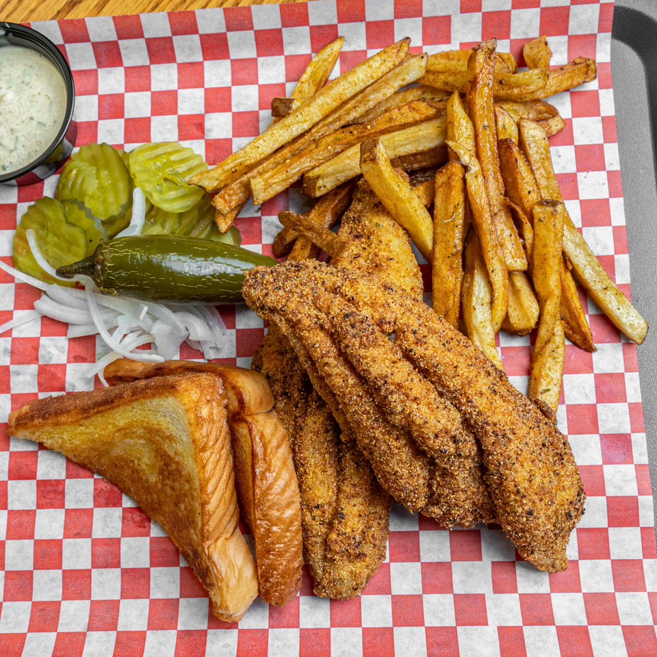 Fried catfish, french fries, toast, pickles, onions, jalapeño, and dipping sauce on checkered paper.